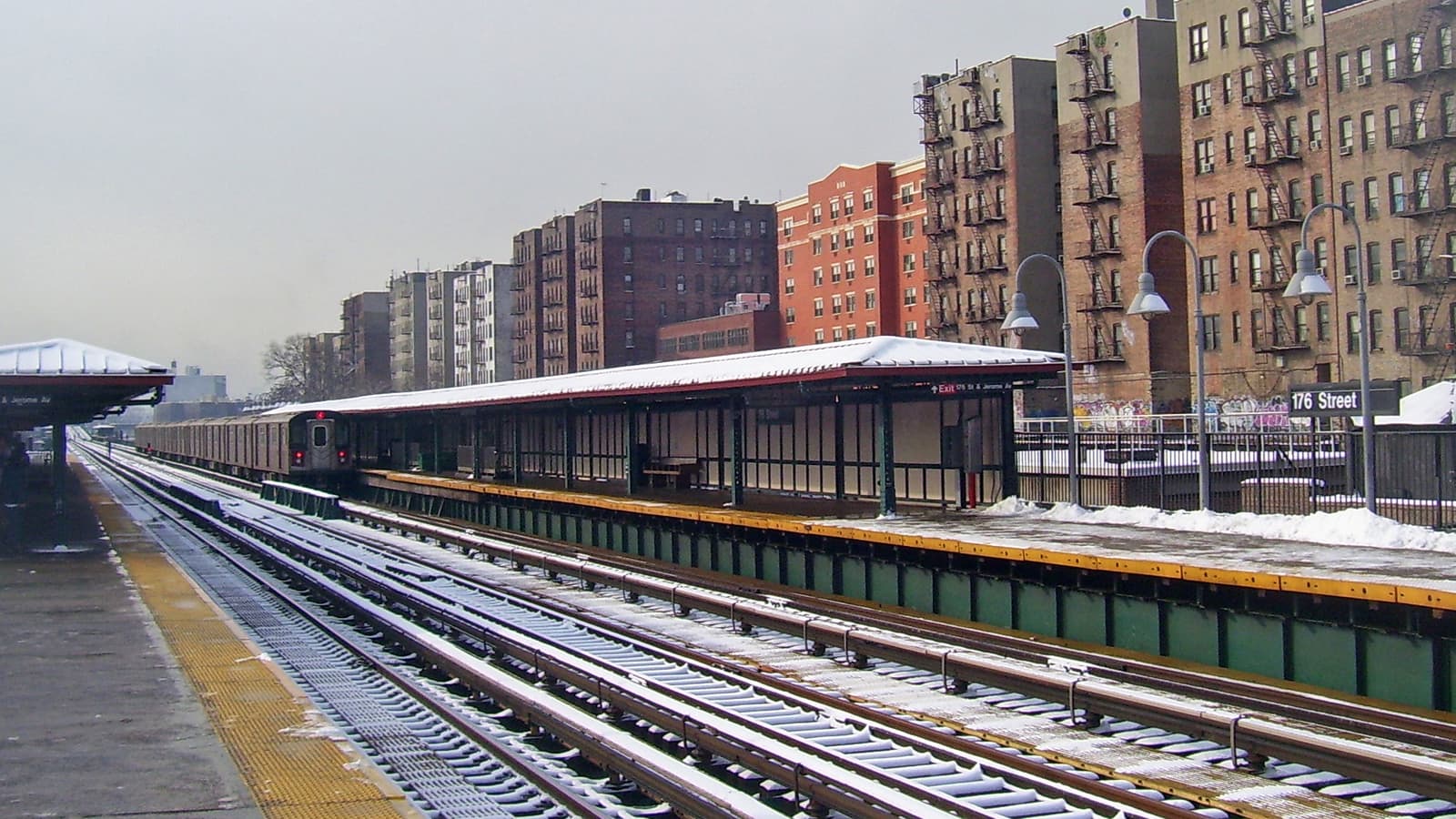 Photograph of a raised subway station in winter, with snow on the platform roofs and tracks, a train pulling away on the far track, a sign reading 176 Street, and tall brick apartment buildings with fire escapes along the right
