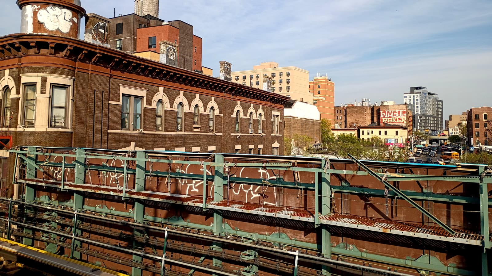 Photograph from a raised subway platform of a rust-brown steel track structure with green railings and graffiti, an old brick building with arched windows and a copper-domed corner tower, and taller apartment buildings and a busy avenue in the distance under a blue sky