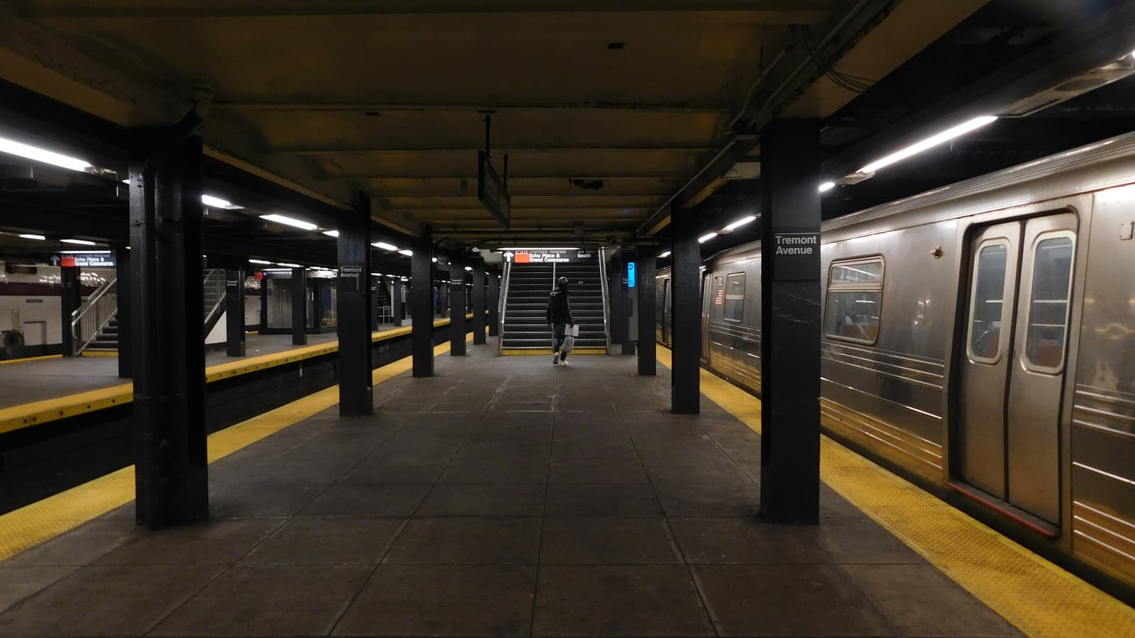 Photograph of an underground subway platform with black steel columns, yellow platform edges, a silver train with closed doors at right, signs reading Tremont Avenue on the columns, and one person walking away toward a staircase in the distance