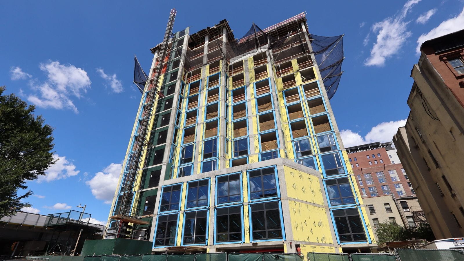 Photograph looking up at a tall concrete apartment building under construction: its lower floors have large dark windows framed in blue tape and yellow sheathing, the floors above are open or partly glazed, black netting hangs around the top, a construction hoist tower runs up the left side and green fencing lines the street, with a railway viaduct at the left edge and older apartment buildings at the right