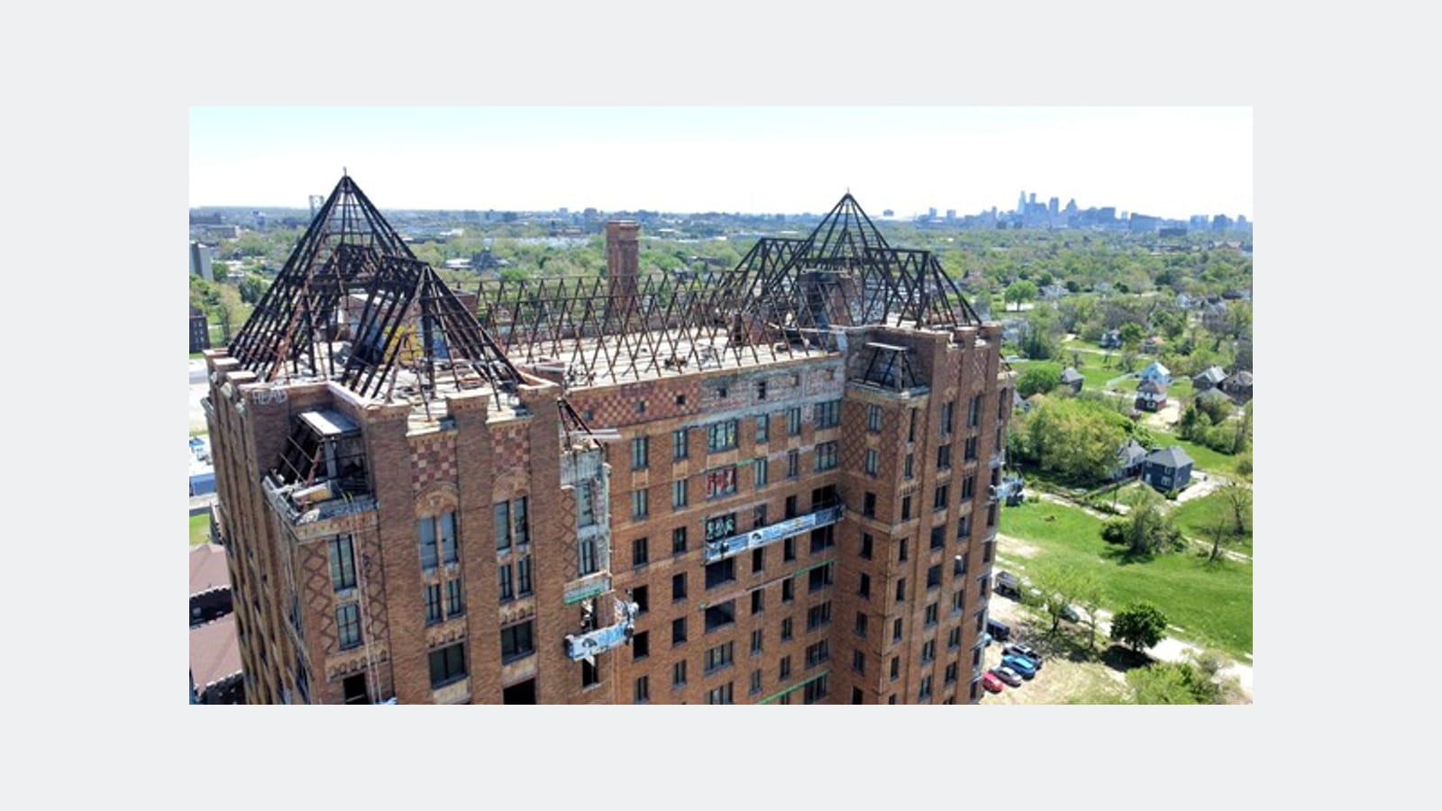 Aerial photograph of the top floors of a brick tower with the bare steel frames of its pointed roofs exposed, a work platform hanging on one side and neighborhoods and a distant skyline behind