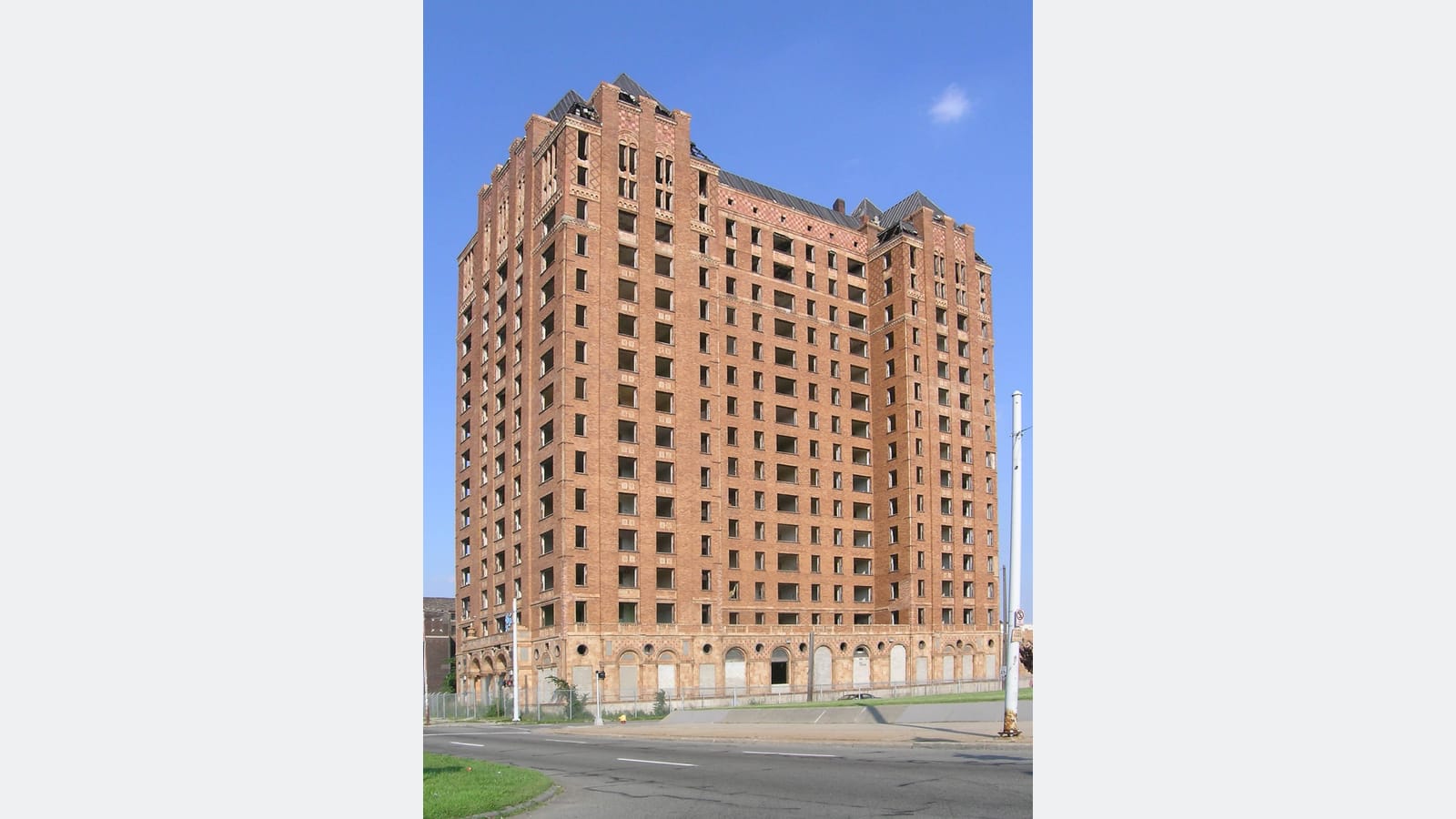 Photograph of a 15-story orange brick tower with empty window openings and steep roofs at its corners, seen from across a street under a blue sky