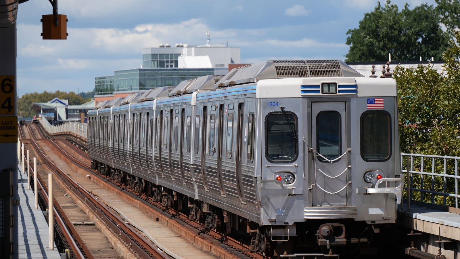 Silver elevated train with a blue stripe leaving a station on tracks above the street, with trees and buildings behind