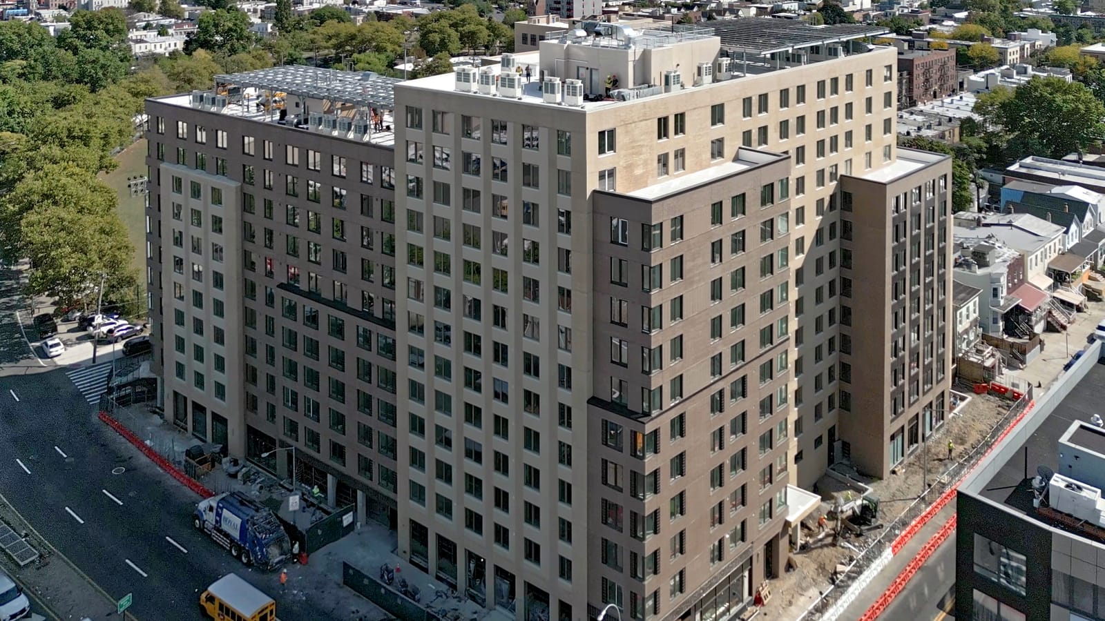 Aerial photograph of a large tan and brown brick building filling the end of a block, with solar panel canopies on two roof levels, construction fencing and orange barriers along the sidewalks, a garbage truck and a school bus on the avenue, and trees and rowhouses beyond