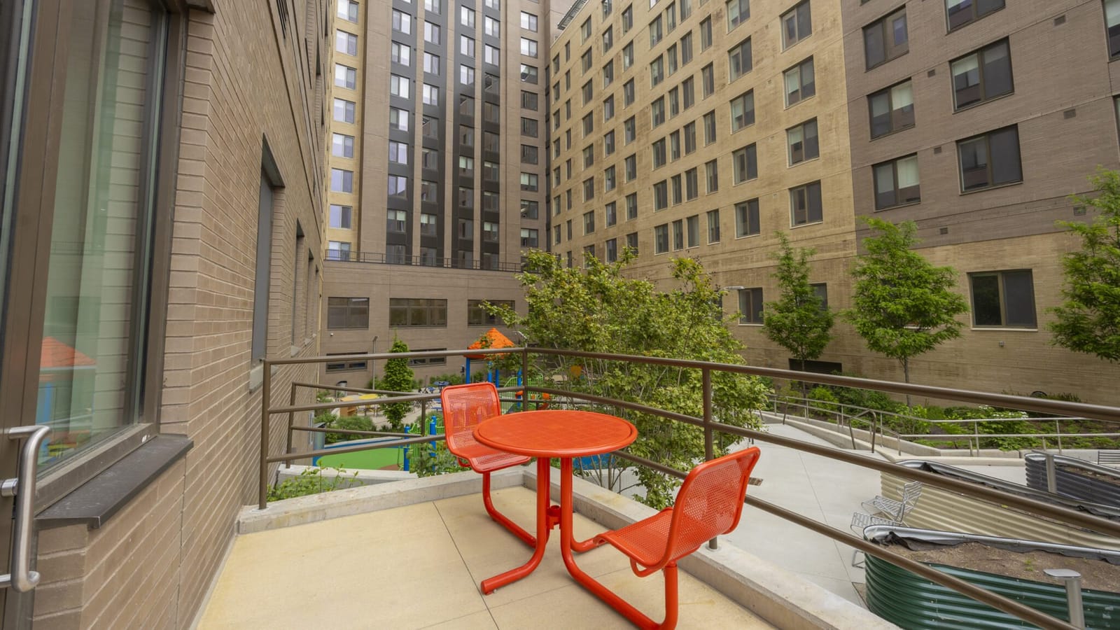Photograph from a terrace with an orange metal table and two chairs, looking over a railing into a courtyard with young trees, a ramp and a playground with green and orange equipment, enclosed by the tan and brown brick wings of the building