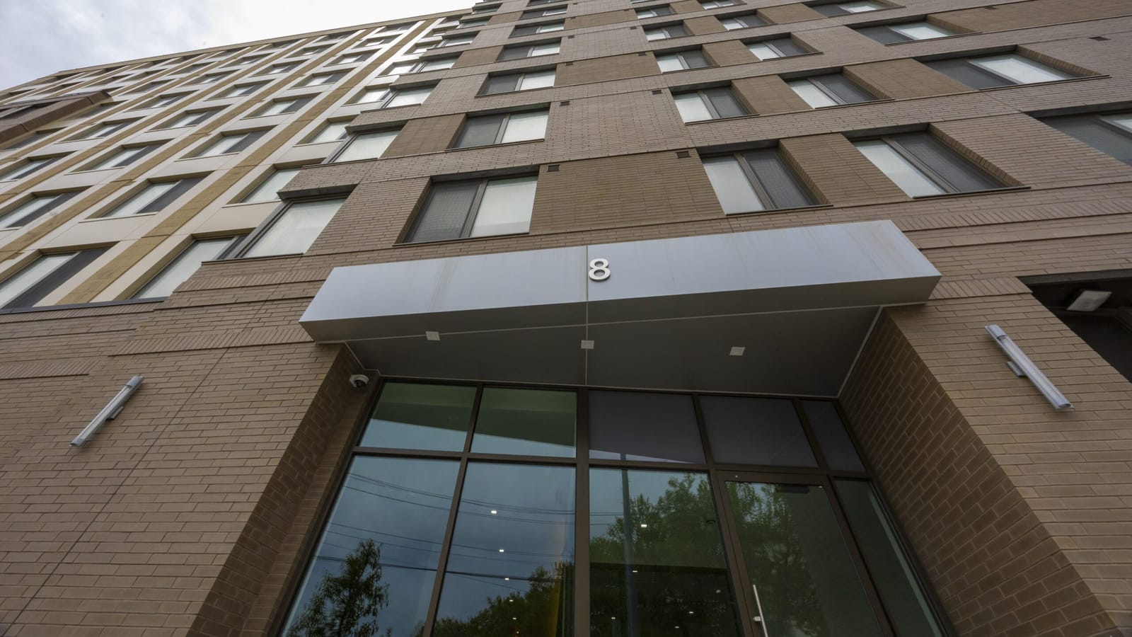 Photograph looking up from the sidewalk at a tan and brown brick building, with a grey metal canopy carrying the number 8 over a glass entrance and rows of windows rising above it