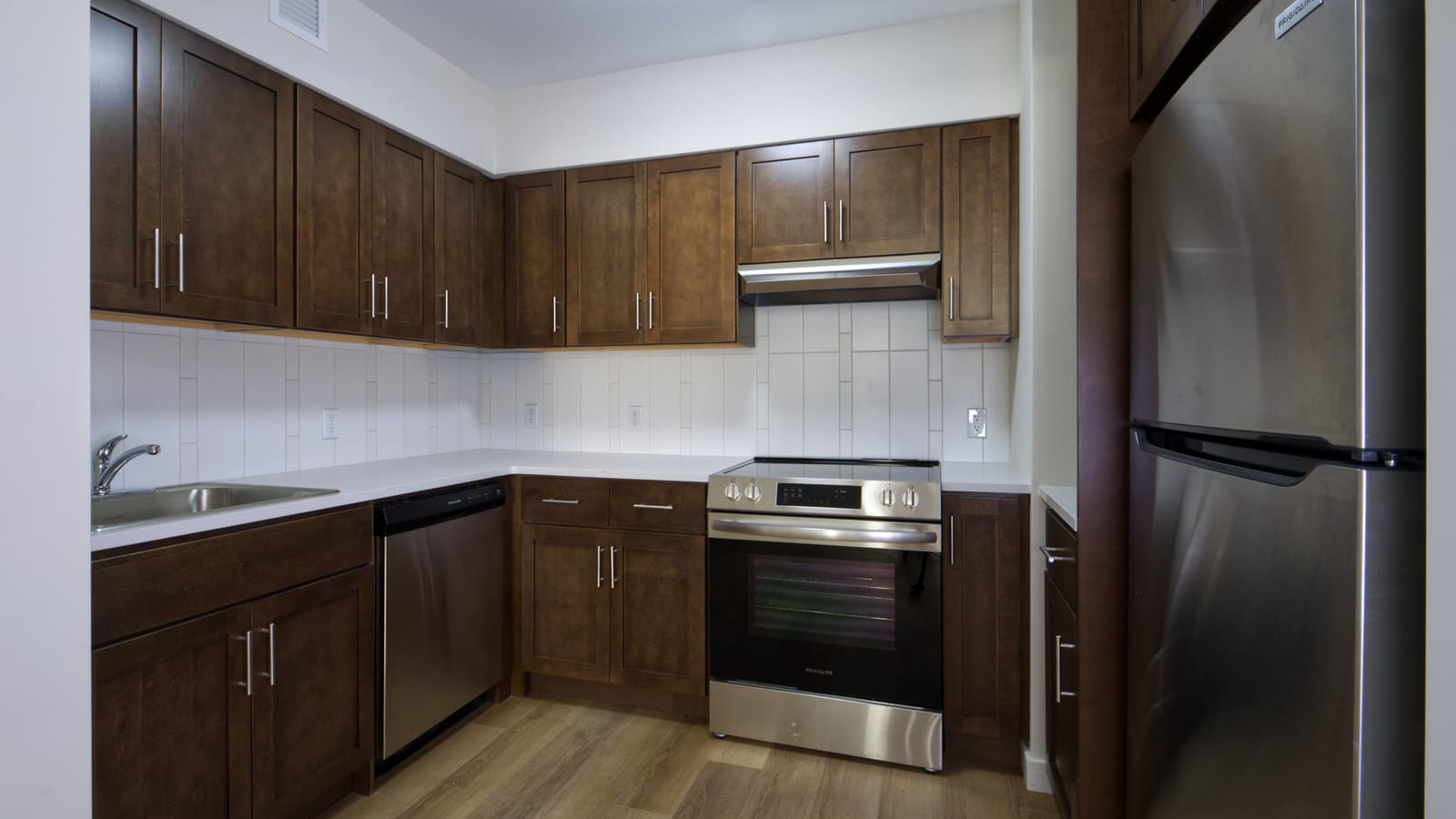 Photograph of an empty kitchen with dark wood cabinets, white countertops and a white tile backsplash, a stainless-steel sink, dishwasher, smooth-top electric range, range hood and refrigerator, and wood-look plank flooring