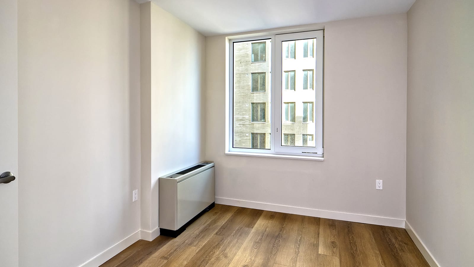 Photograph of an empty room with white walls, wood-look plank flooring, a two-panel window facing another brick wing of the building, and a floor-standing heating and cooling unit beside the window wall