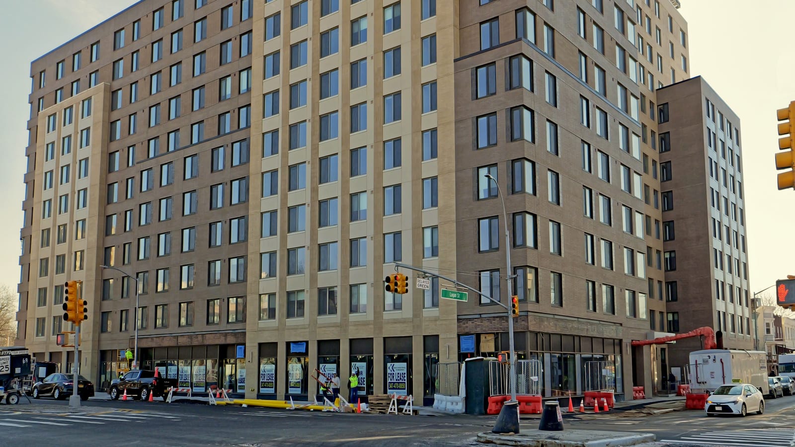 Photograph of the lower floors of a tan and brown brick building at a street corner with a Logan St sign, ground-floor storefront windows carrying FOR LEASE signs, construction barriers and cones on the sidewalk, workers in yellow vests and parked cars