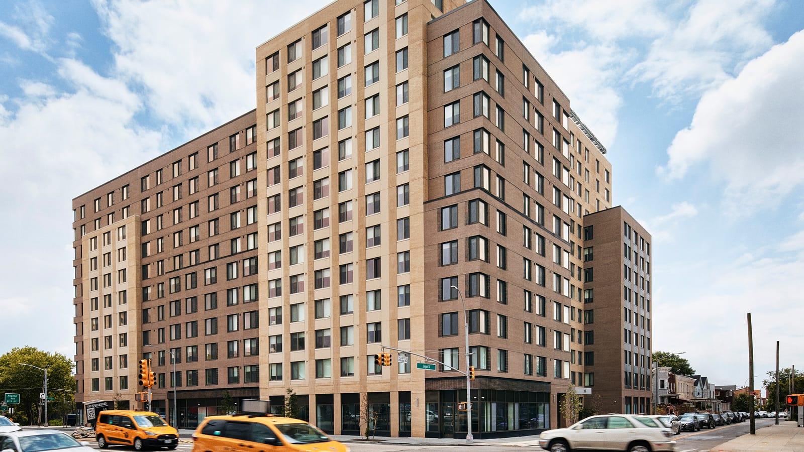 Photograph of a tall apartment building in tan and brown brick at a street corner, with glass storefronts along the ground floor, a street sign reading Logan St on a traffic signal, and two yellow taxis and other cars passing in the foreground