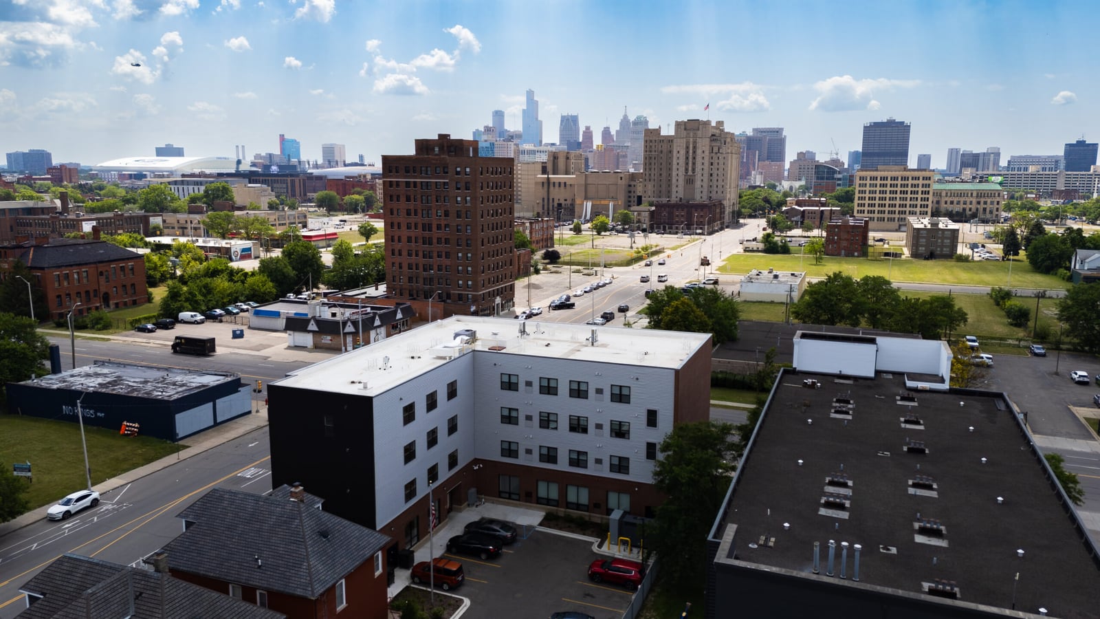 Aerial photograph of an L-shaped four-story building with white and dark siding and a parking lot behind it, with a wide avenue and the downtown Detroit skyline beyond