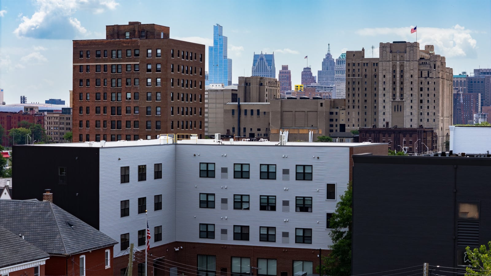 Photograph of the upper floors of a white-sided four-story apartment building with rows of windows, in front of older brick towers and downtown skyscrapers