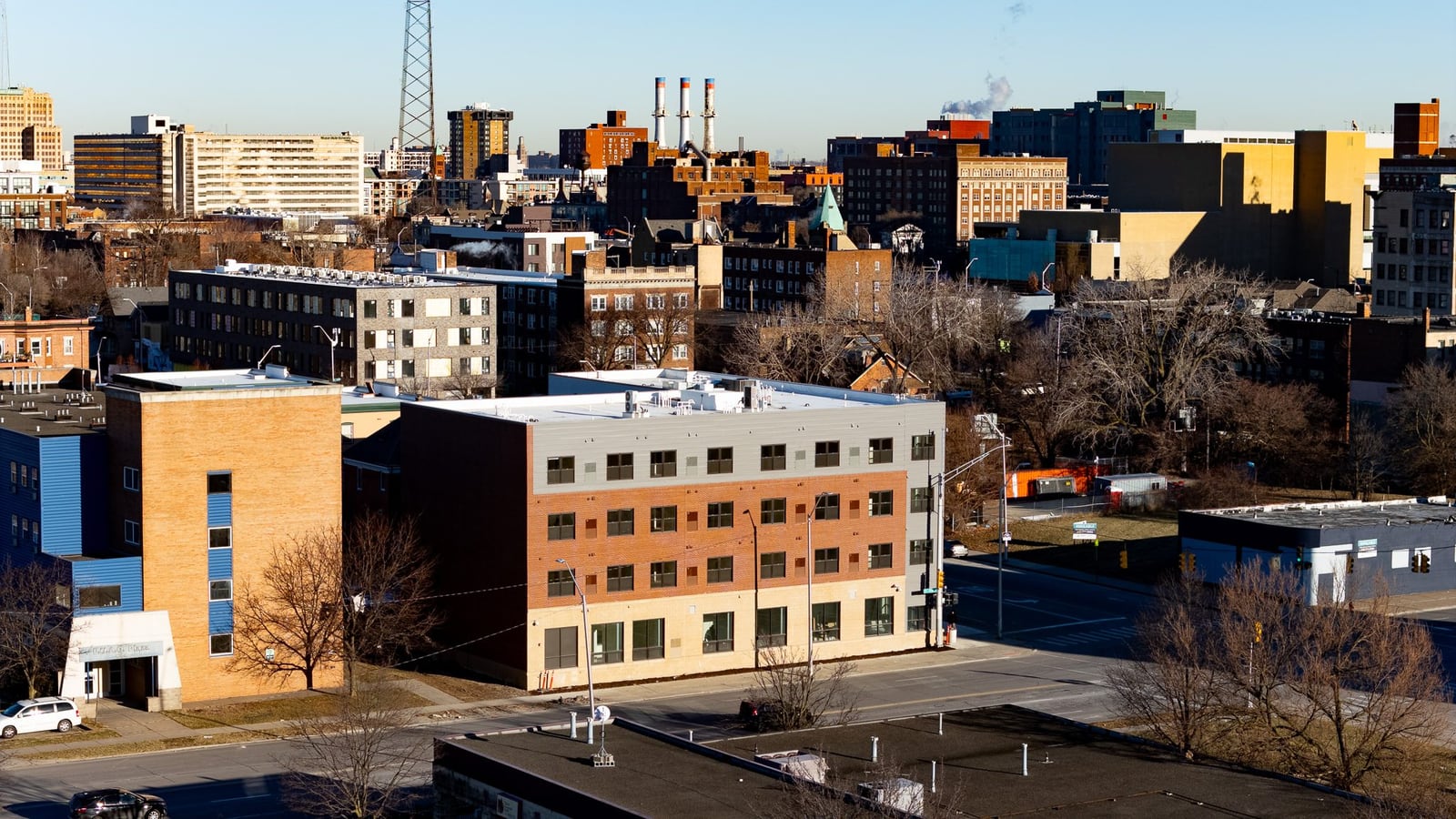 Aerial photograph of a four-story apartment building with a gray top floor, red brick middle floors and a pale ground floor with large windows, beside a tan brick building, with Midtown buildings behind