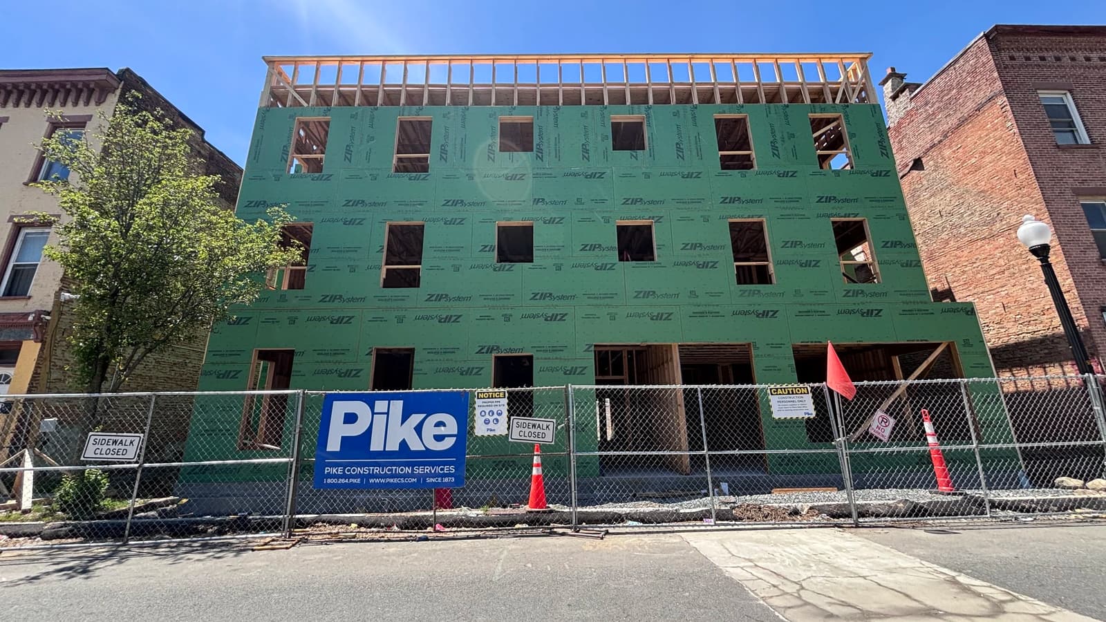A three-story wood-framed building wrapped in green sheathing behind a construction fence with a Pike Construction Services banner, between two brick buildings