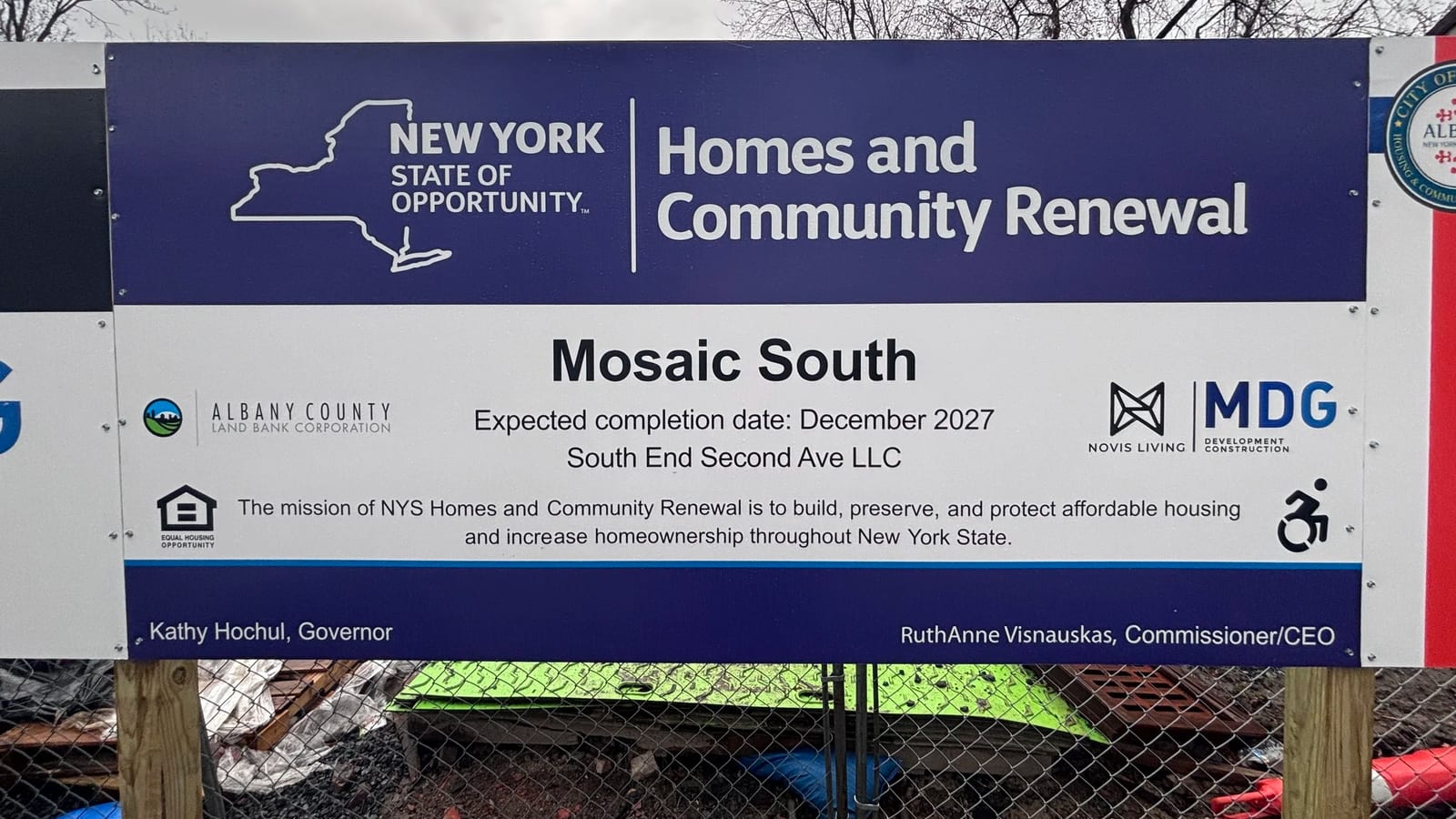 Blue and white construction sign reading New York State Homes and Community Renewal, Mosaic South, expected completion date December 2027, South End Second Ave LLC, with Albany County Land Bank, Novis Living and MDG logos