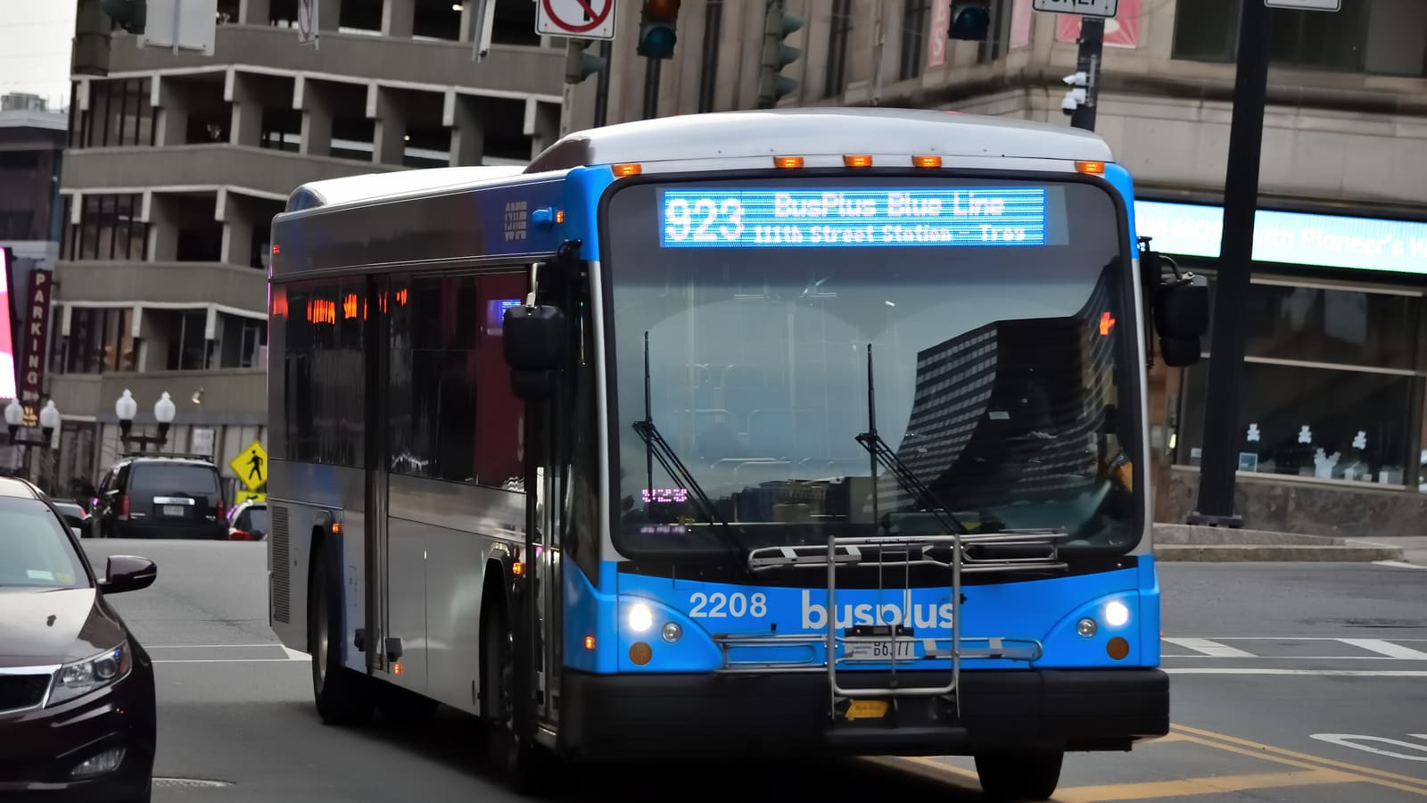 A blue and silver bus with a sign reading 923 BusPlus Blue Line at a downtown intersection
