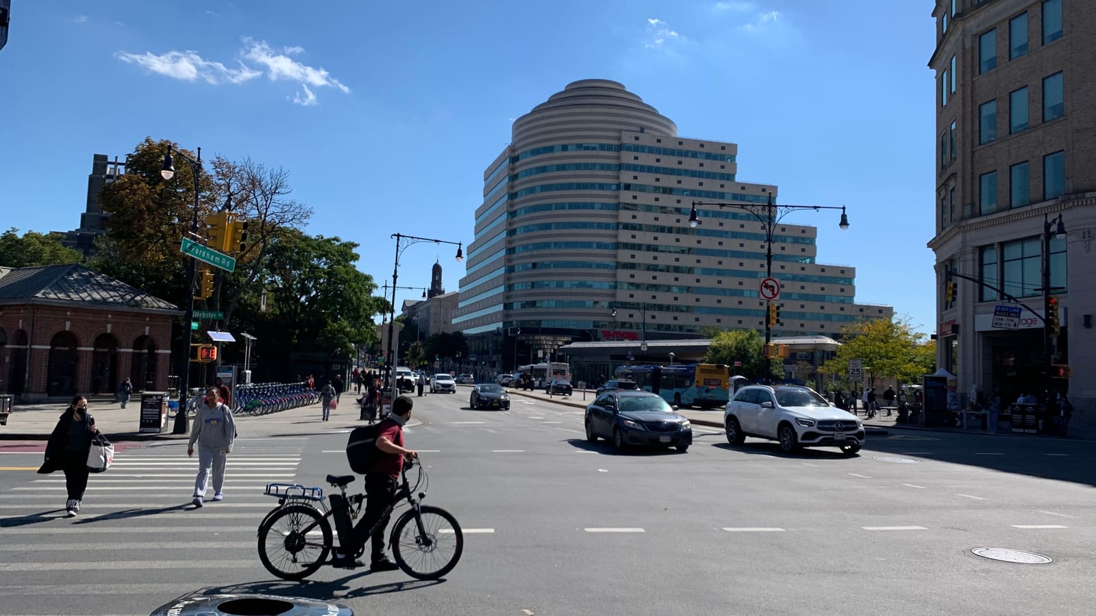 A stepped, round-fronted office tower behind a wide street intersection with cars, a bus and people crossing