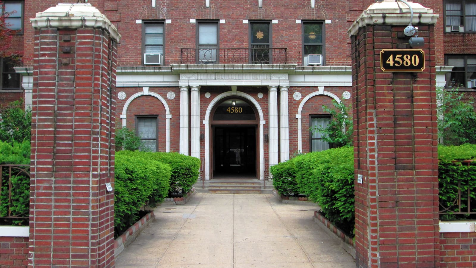 The arched entrance of a red-brick apartment building behind brick gate posts and hedges