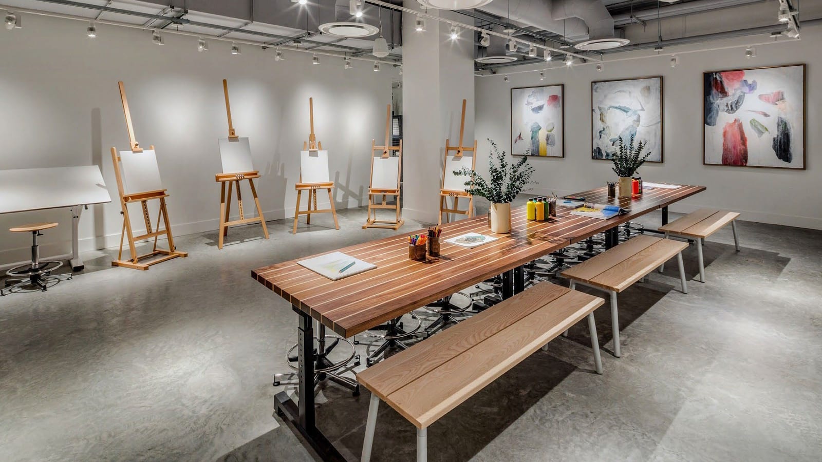 An art room with five wooden easels holding blank canvases, a long wooden table with benches, paint bottles, plants and three abstract paintings on a white wall