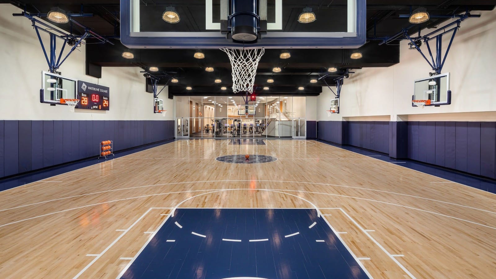 An indoor basketball court with a wood floor, navy wall pads, six hoops and a scoreboard reading Waterline Square, with a glass wall to a gym at the far end