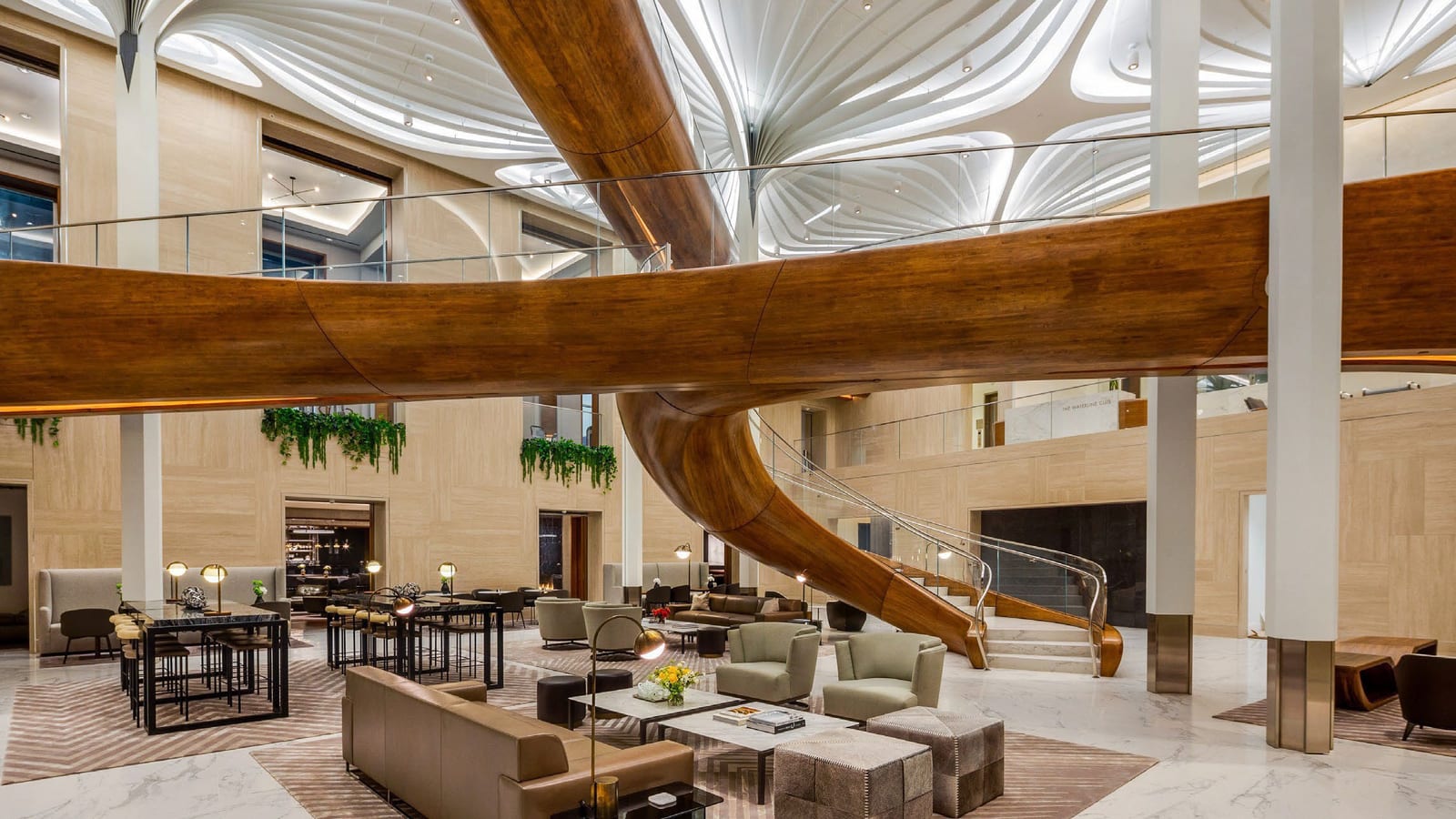 A tall lounge with a curving wooden slide that loops under a white sculpted ceiling, a marble floor, sofas, armchairs and high tables