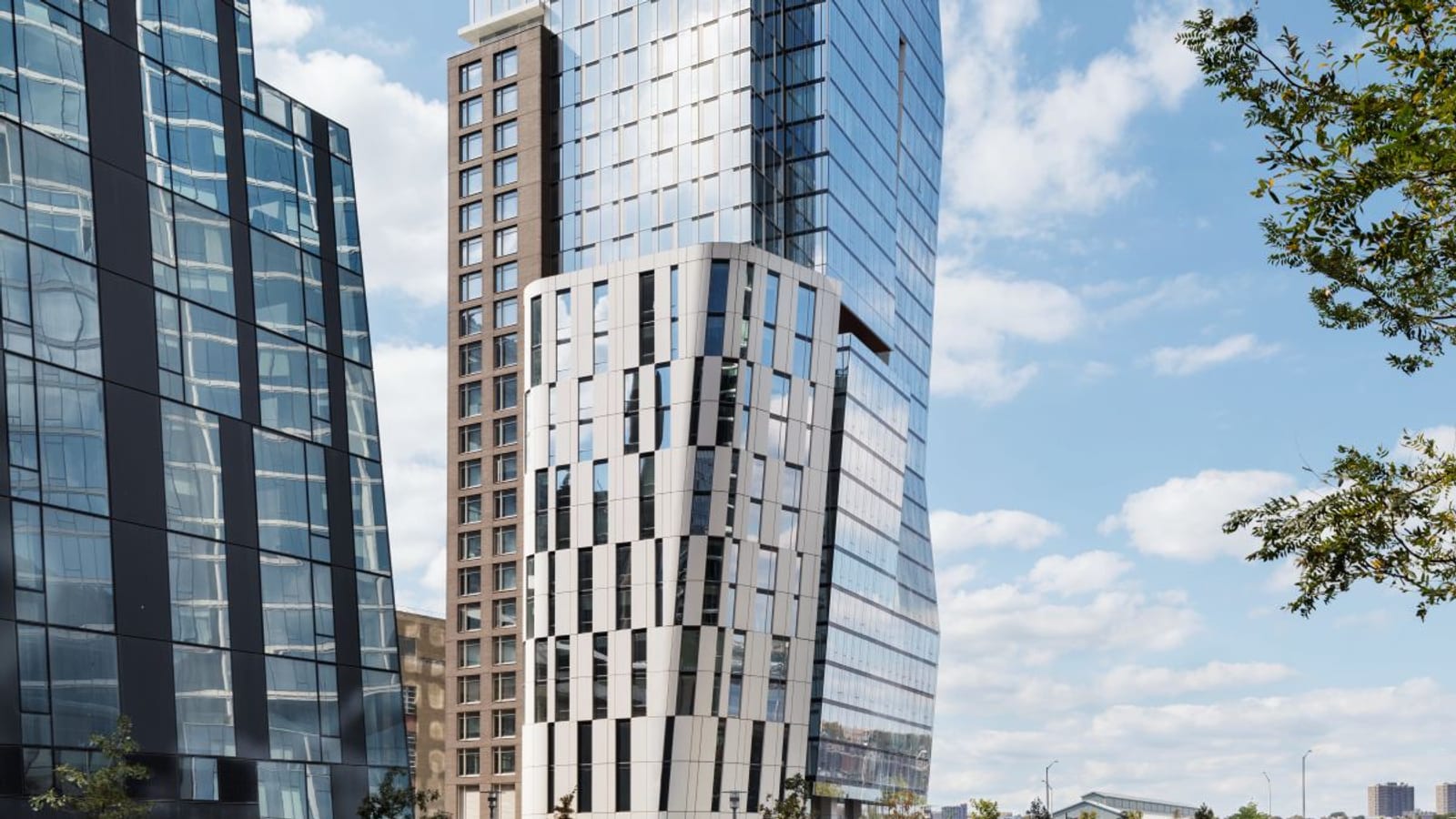 A slim glass tower rising behind a curved base clad in white panels with narrow black windows, next to a brown brick wing, with a dark glass tower at left and trees and blue sky at right