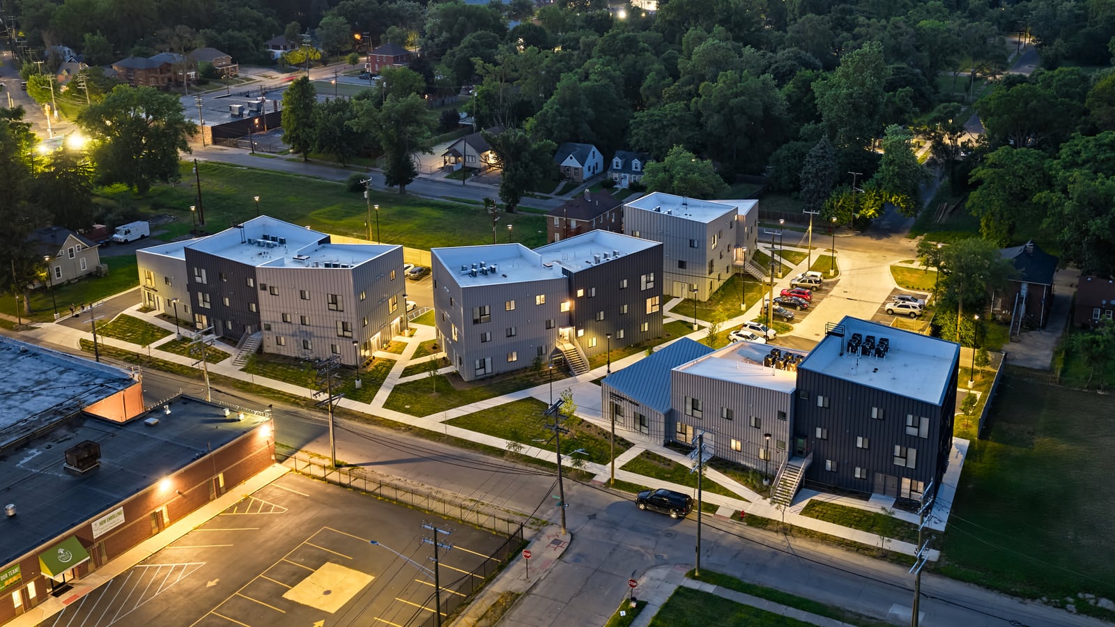 Aerial photograph at sunset of lit apartment buildings with white roofs, outside stairs and paths between them, surrounded by houses and trees