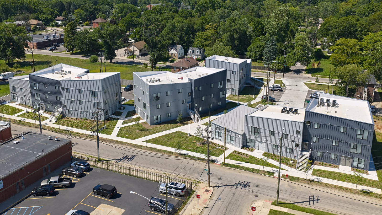 Aerial photograph of gray metal-clad apartment buildings of two and three stories with white roofs, linked by paths and lawns, beside a street and a parking lot, with trees to the horizon
