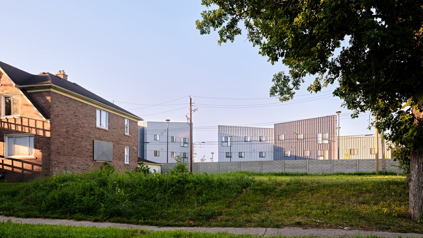 Photograph across a grassy lot of gray metal-clad apartment buildings behind a block wall, with a brick house at left and a large tree at right