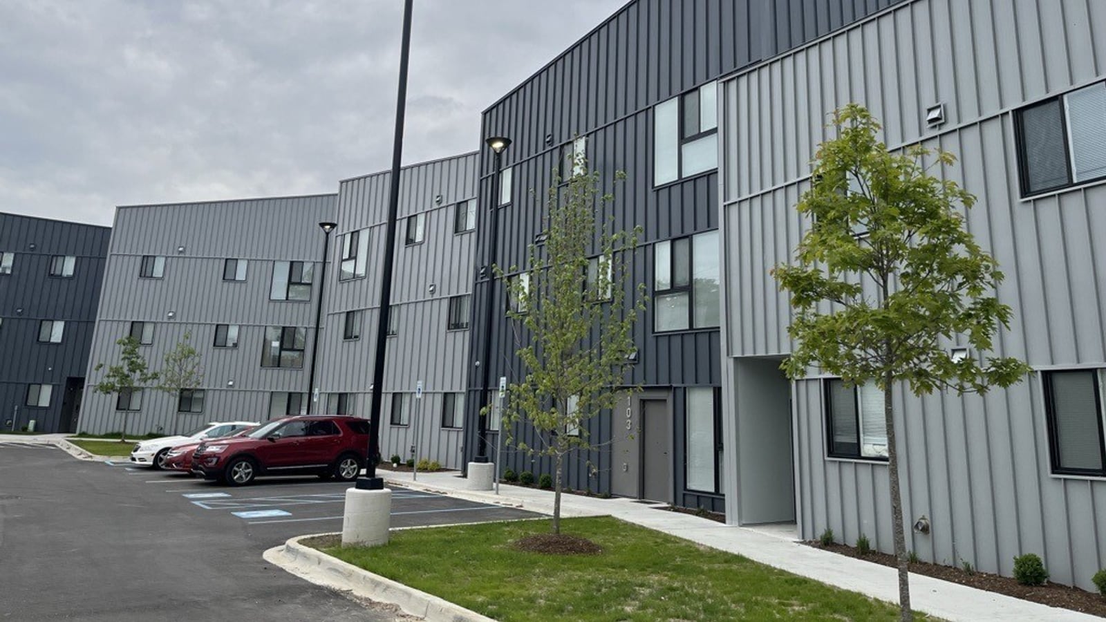 Photograph of three-story apartment buildings clad in light and dark gray metal panels beside a parking lot with marked accessible spaces, lamp posts and young trees