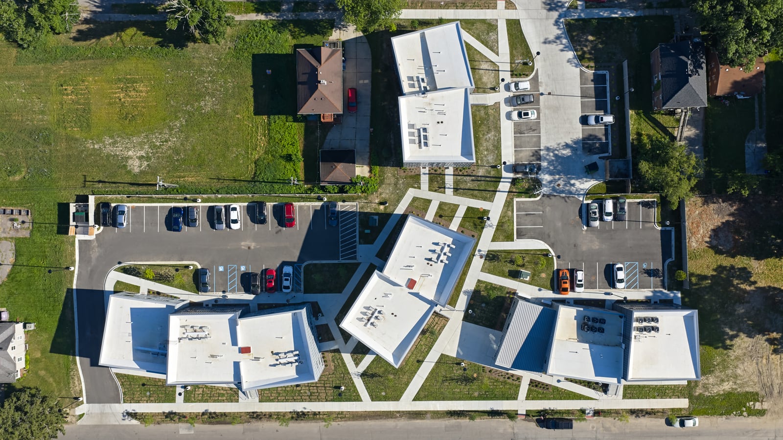 Photograph taken straight down on four white-roofed apartment buildings set at angles between two streets, with two parking lots, diagonal paths and lawns