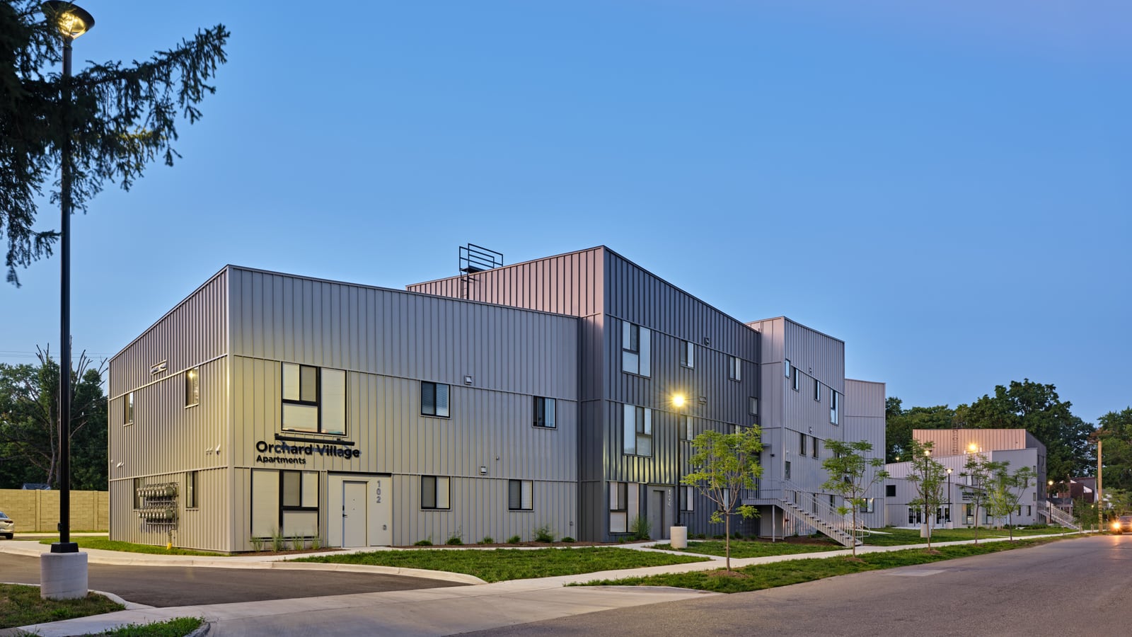 Photograph at dusk of a gray metal-clad apartment building of two and three stories lettered Orchard Village Apartments, with lit lamps, young trees and more buildings behind it