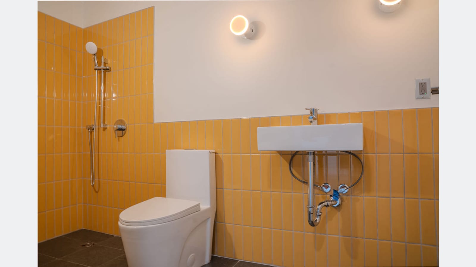A bathroom tiled in yellow to shoulder height with an open shower and no tub, a floor drain, a toilet and a wall-hung sink