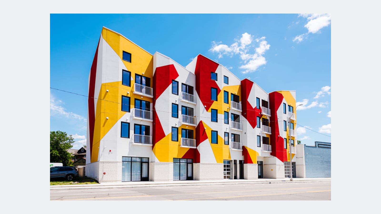 Photograph of an apartment building with a front painted in large red, yellow and white angular shapes, a sawtooth roofline, a glass shop level and four rows of windows above it, under a blue sky