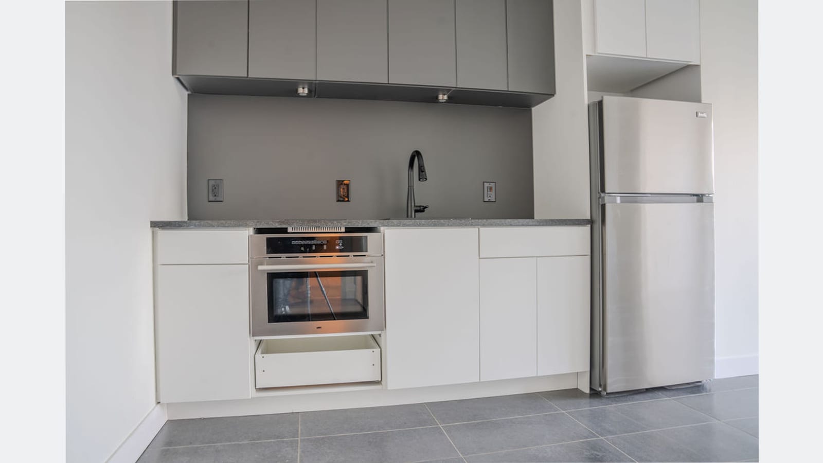 A kitchen along one wall with gray upper cabinets, white lower cabinets, a built-in stainless steel oven, a sink and a stainless steel refrigerator, on a gray tile floor