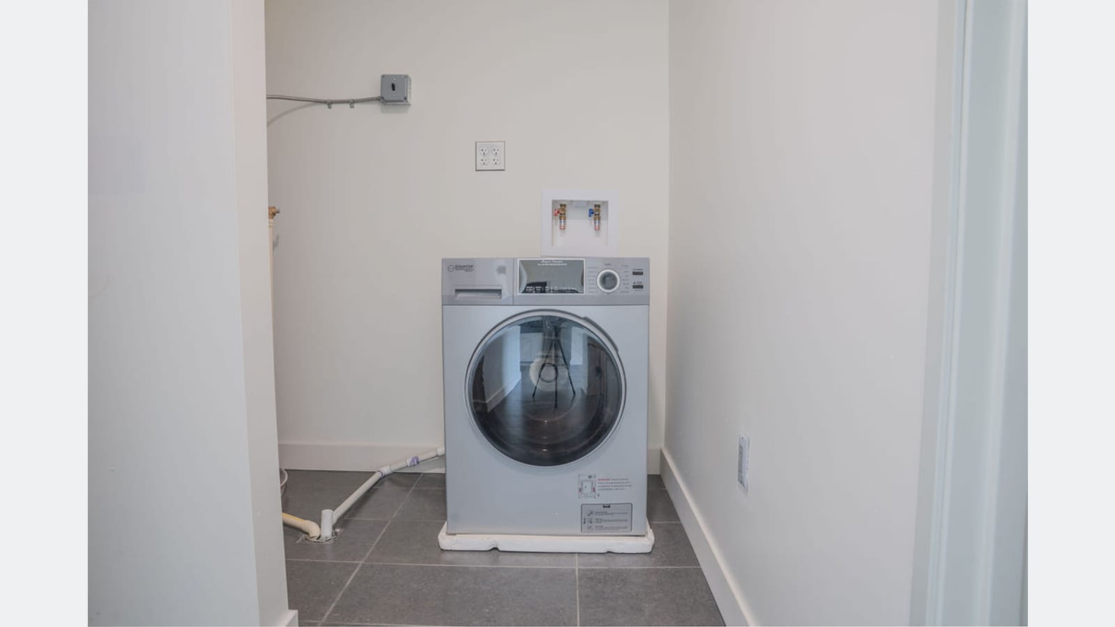 A single front-loading laundry machine standing in a closet with water hookups on the wall above it