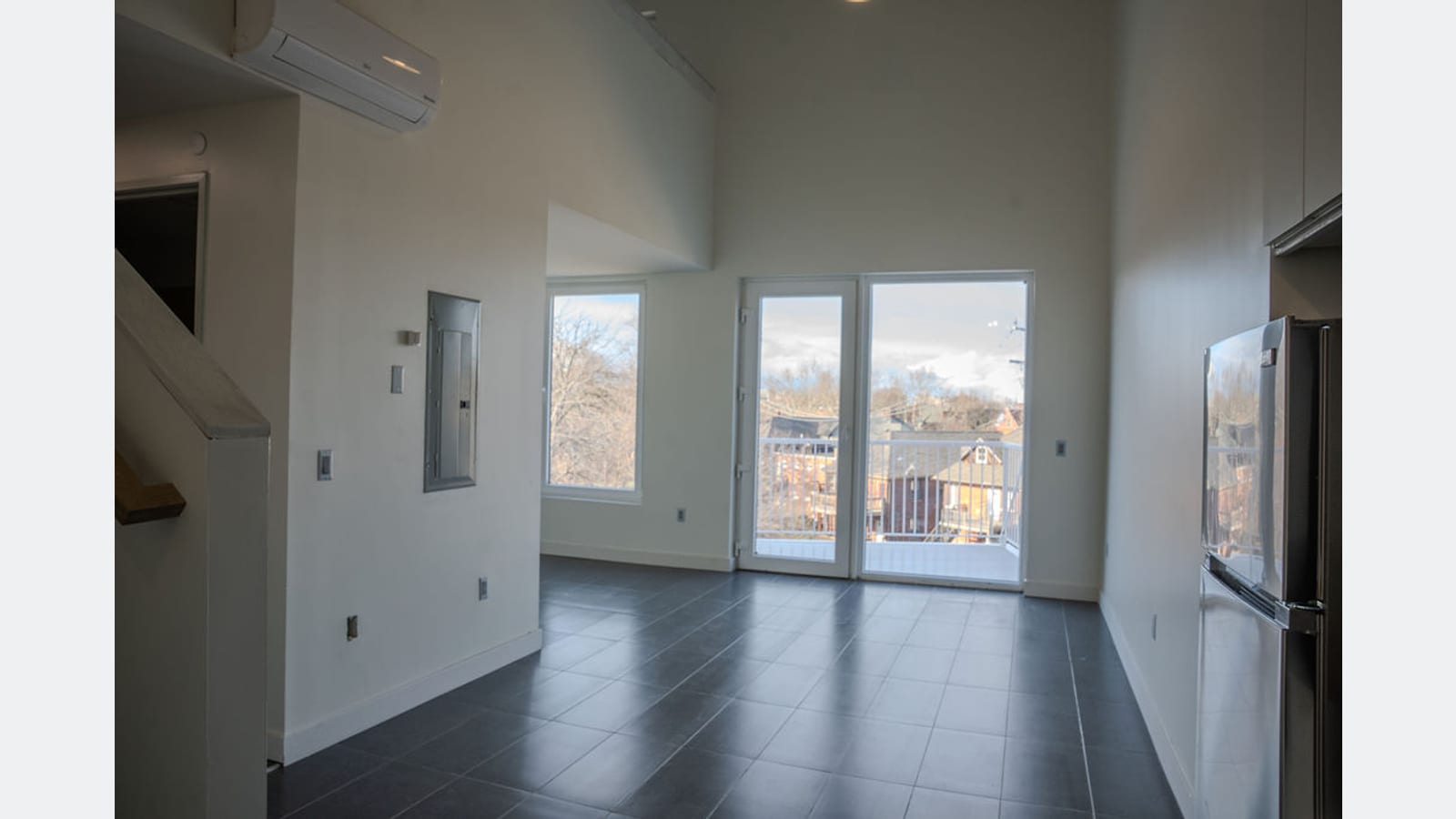 A two-floor apartment seen from its kitchen: a high living room with a gray tile floor, glass doors to a balcony, a wall-mounted air unit near the ceiling and the foot of a staircase at the left