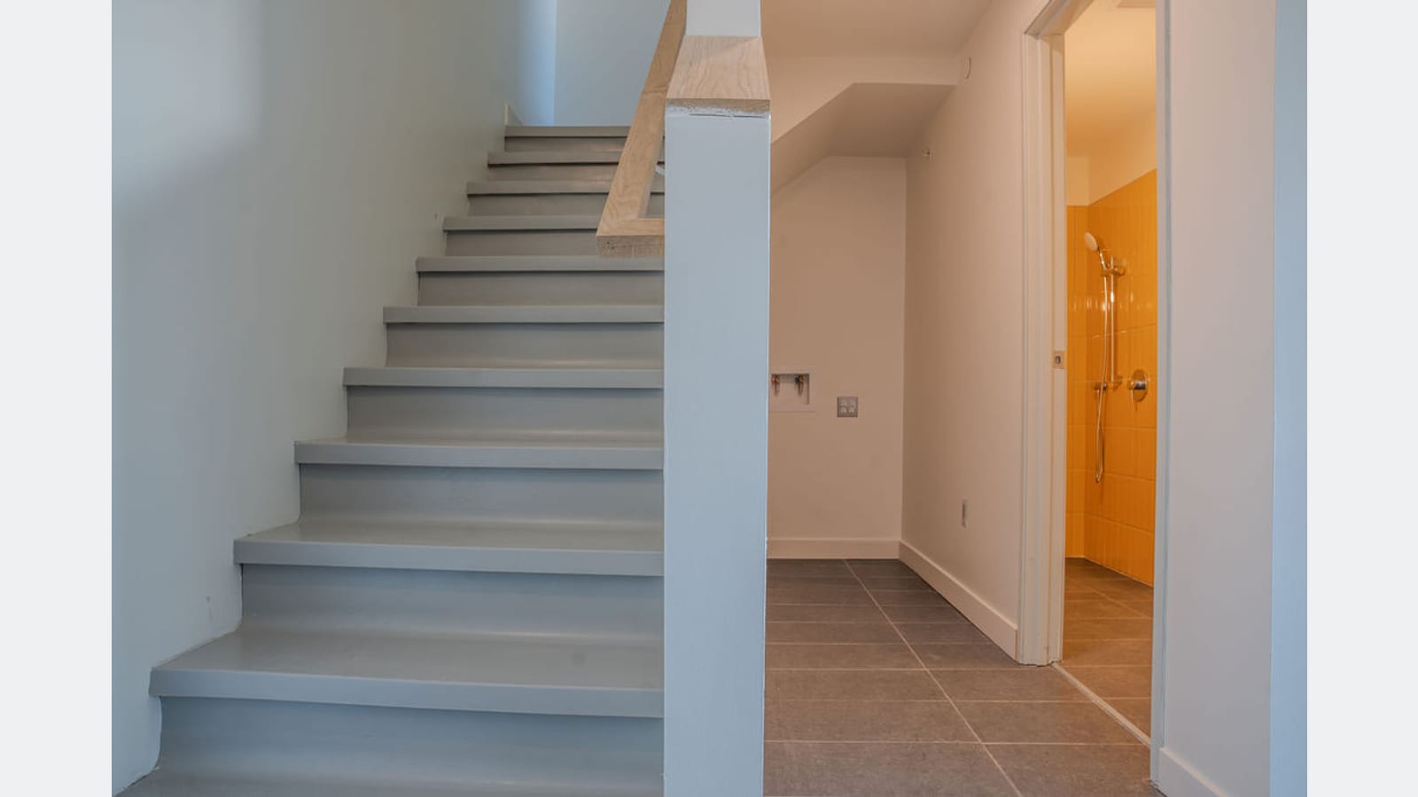A staircase inside an apartment with a pale wood handrail, beside a hall with laundry hookups and a doorway to a yellow-tiled bathroom