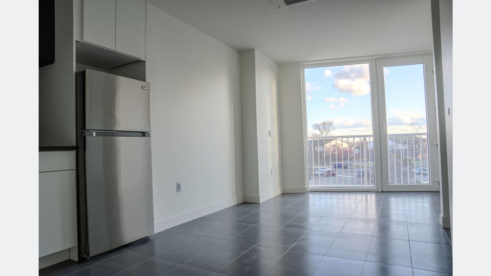 An empty studio apartment with a gray tile floor: a stainless steel refrigerator and cabinets at the left and glass doors to a railed balcony at the far end