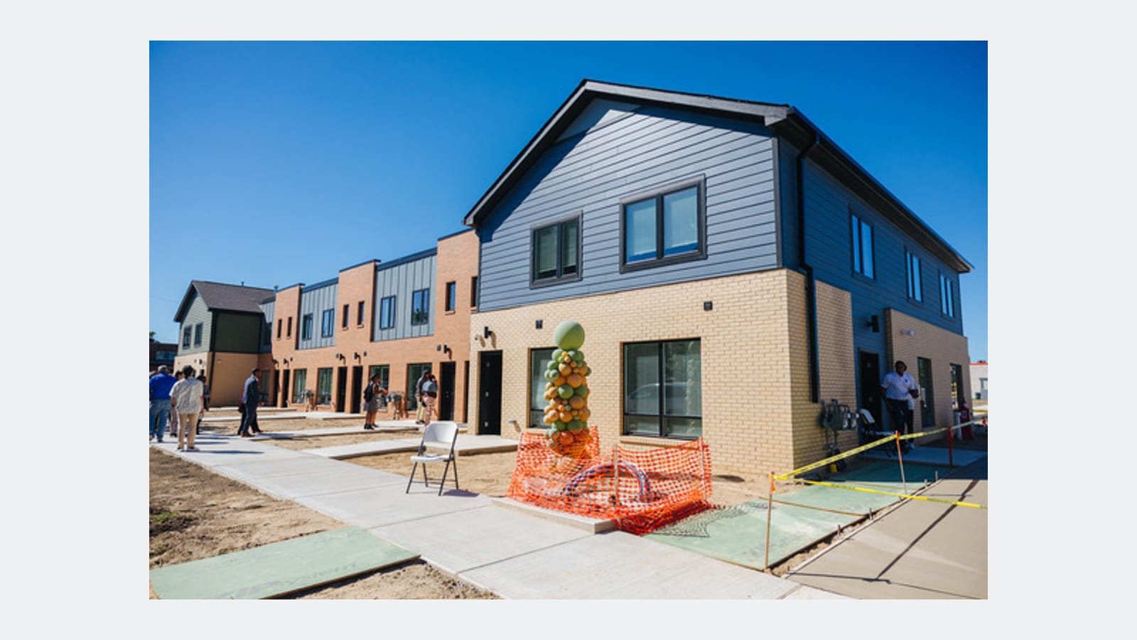 Photograph of a row of two-story townhomes with blue-gray siding above tan and orange brick, a new sidewalk in front and a few people standing near the doors under a clear sky