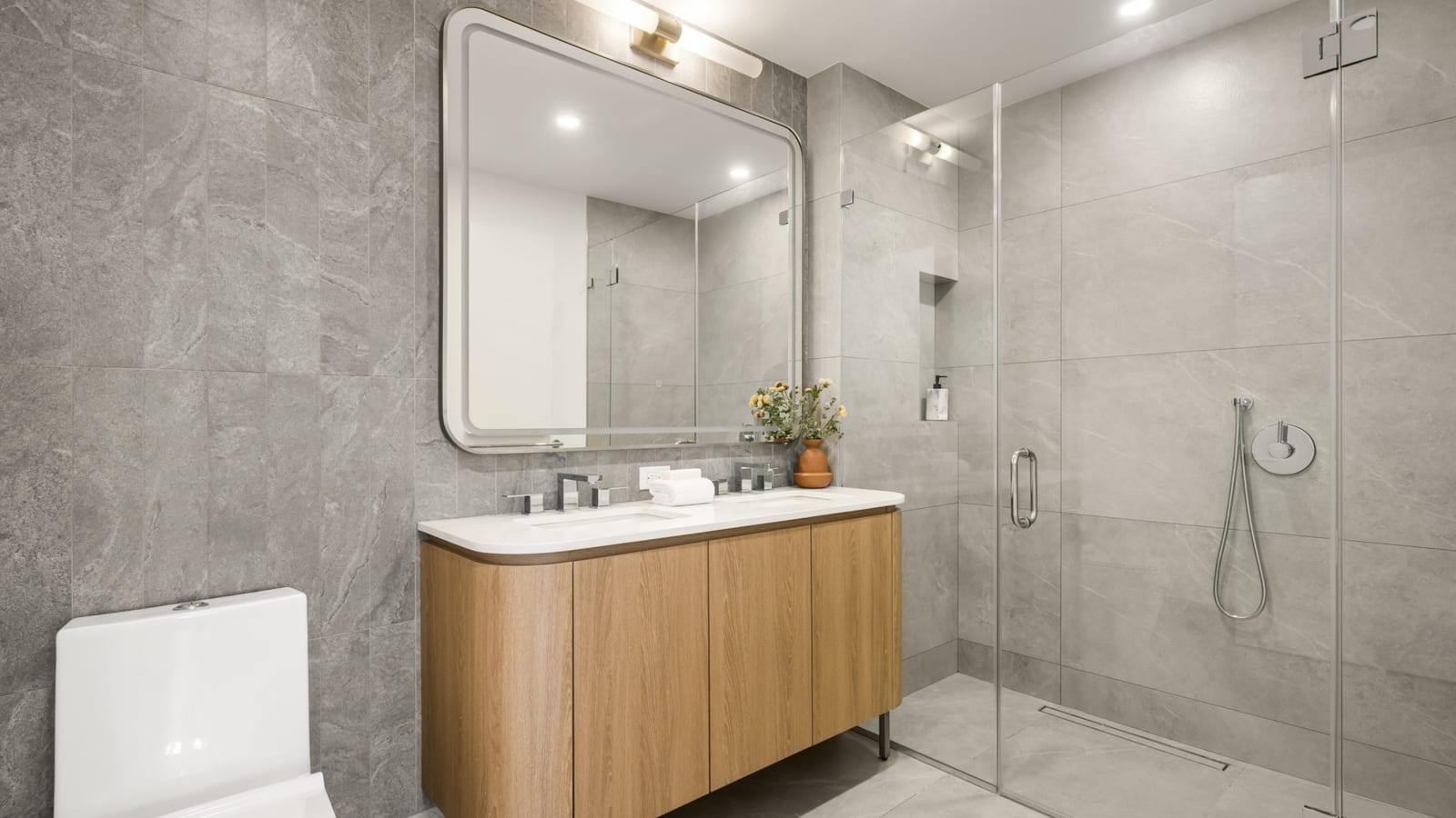 Photograph of a bathroom with large grey stone-look tile on the walls and floor, a wood vanity with a white top and two faucets under a rounded lighted mirror, a white toilet and a glass-enclosed walk-in shower with a handheld sprayer