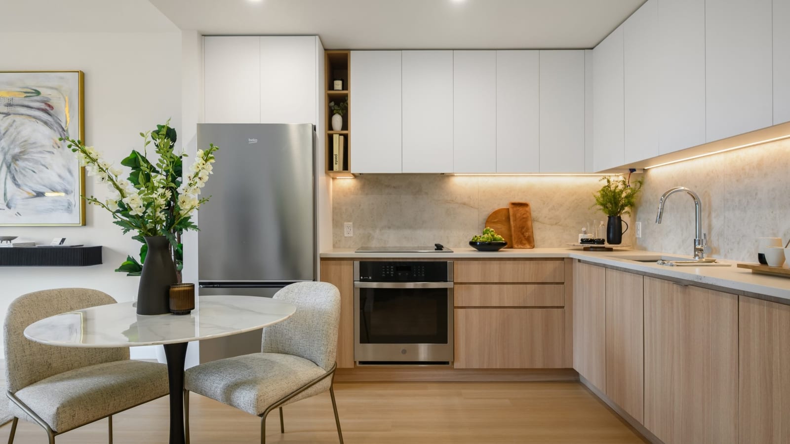 Photograph of a staged apartment kitchen with flat white upper cabinets, wood-tone lower cabinets, a pale stone backsplash lit from under the cabinets, a smooth black cooktop over a built-in stainless-steel wall oven, a stainless-steel refrigerator, a sink with a chrome faucet, and a small round marble table with two upholstered chairs in the foreground