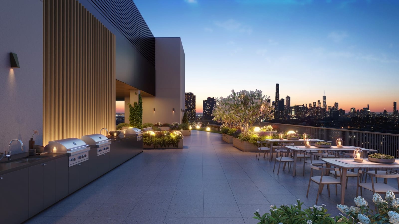 Rendering at dusk of a roof terrace with a paved deck, a counter with a sink and three built-in grills along a wall at left, planters with shrubs and small globe lights, wooden dining tables and chairs along a railing at right, and a skyline of towers on the horizon