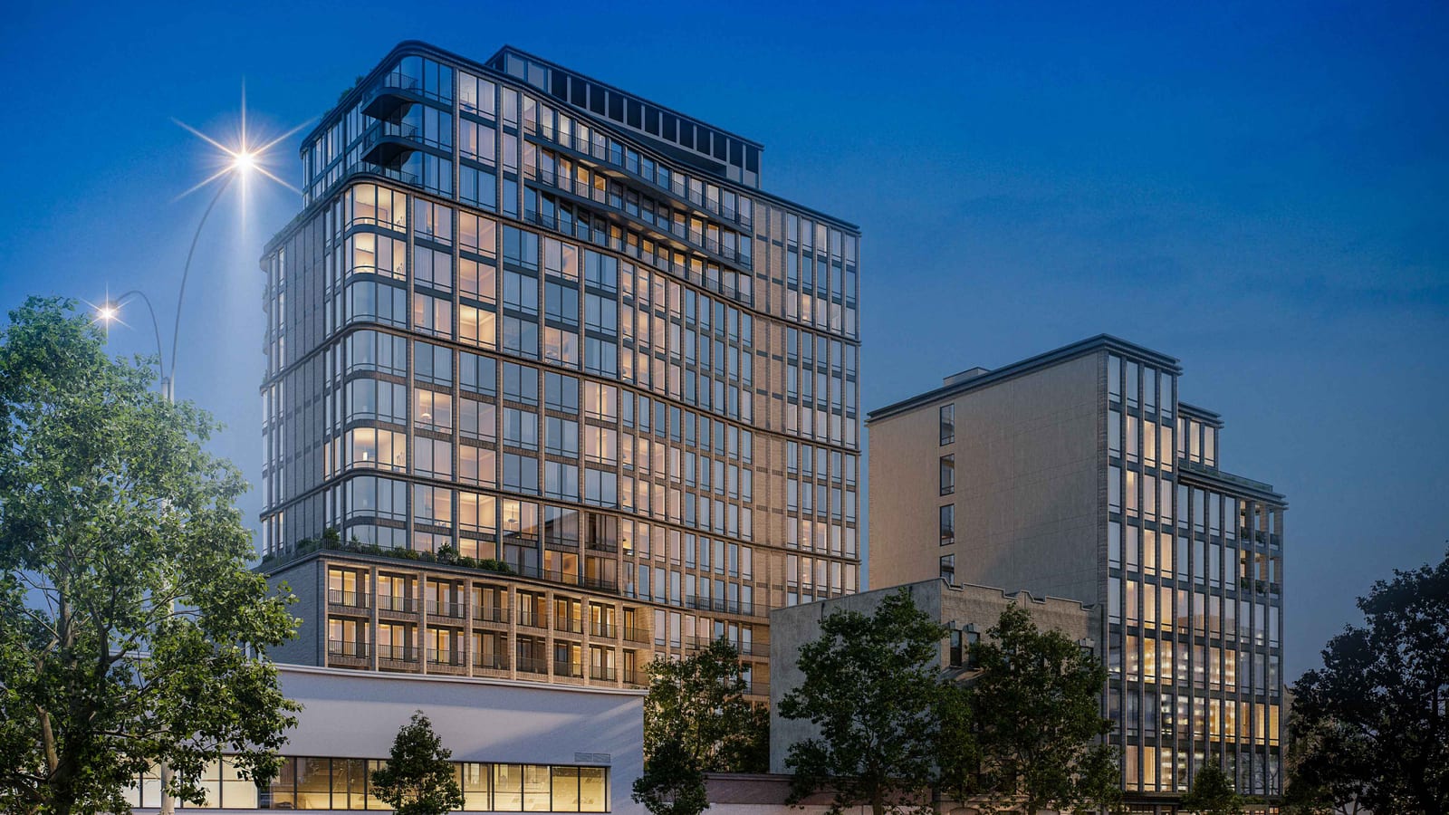 Rendering at dusk of a tall apartment tower with a dark grid of large lit windows, rounded corners, a planted setback above a brick base with balconies and two stacks of curved balconies near the top, standing beside a lower slab-shaped wing of about nine stories, with street trees, a streetlight, a low white building and an older brick building in front