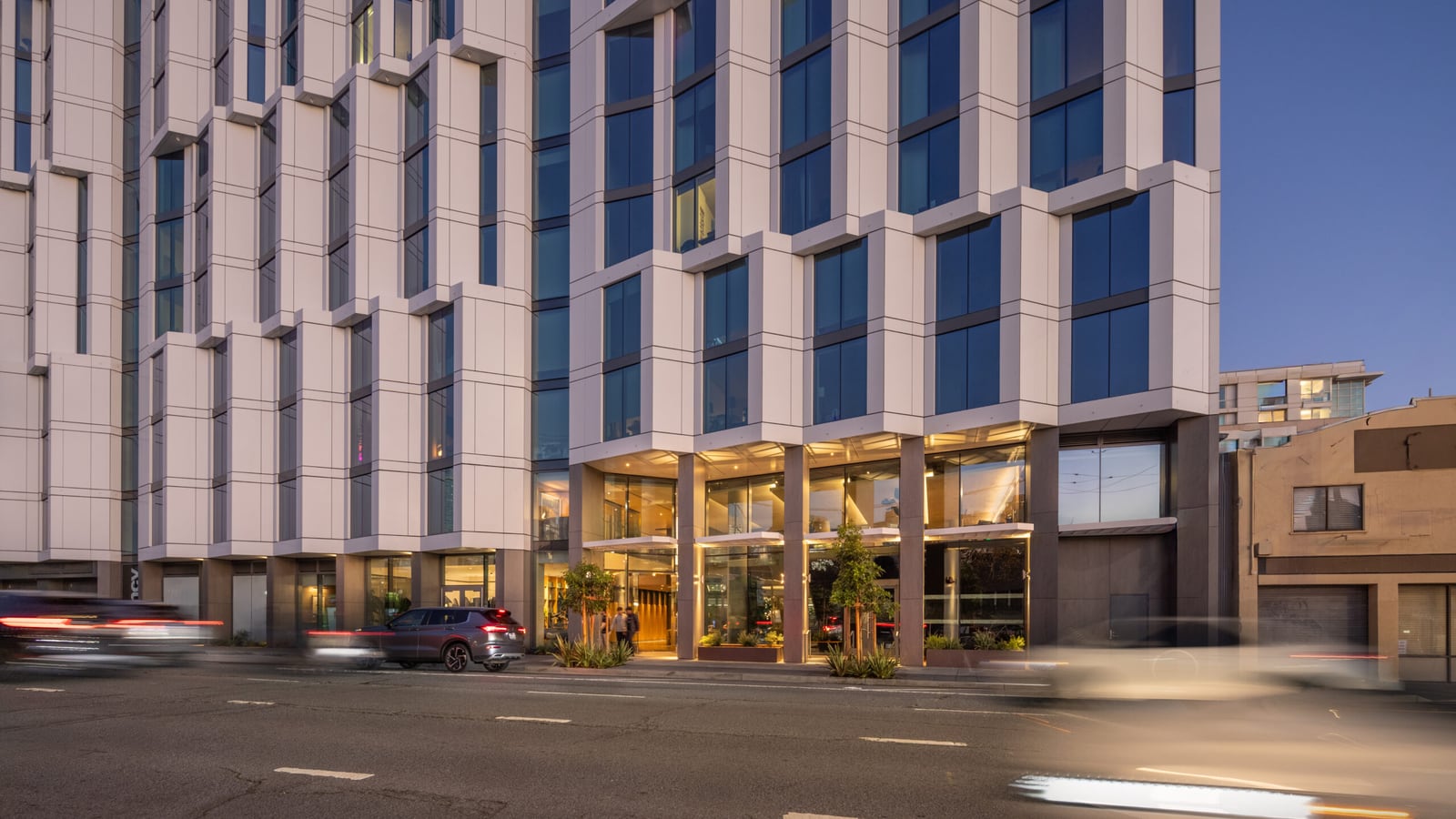 Street-level photograph at dusk of the lower floors of a white apartment building with angled bay windows above a glass-walled lobby, with cars passing on the street