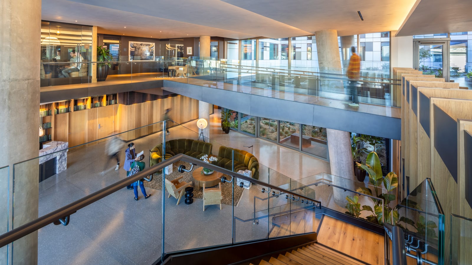 Interior photograph of a two-level lobby seen from a wooden staircase: a seating area with green sofas below, a mezzanine with glass railings and work tables above, and concrete columns