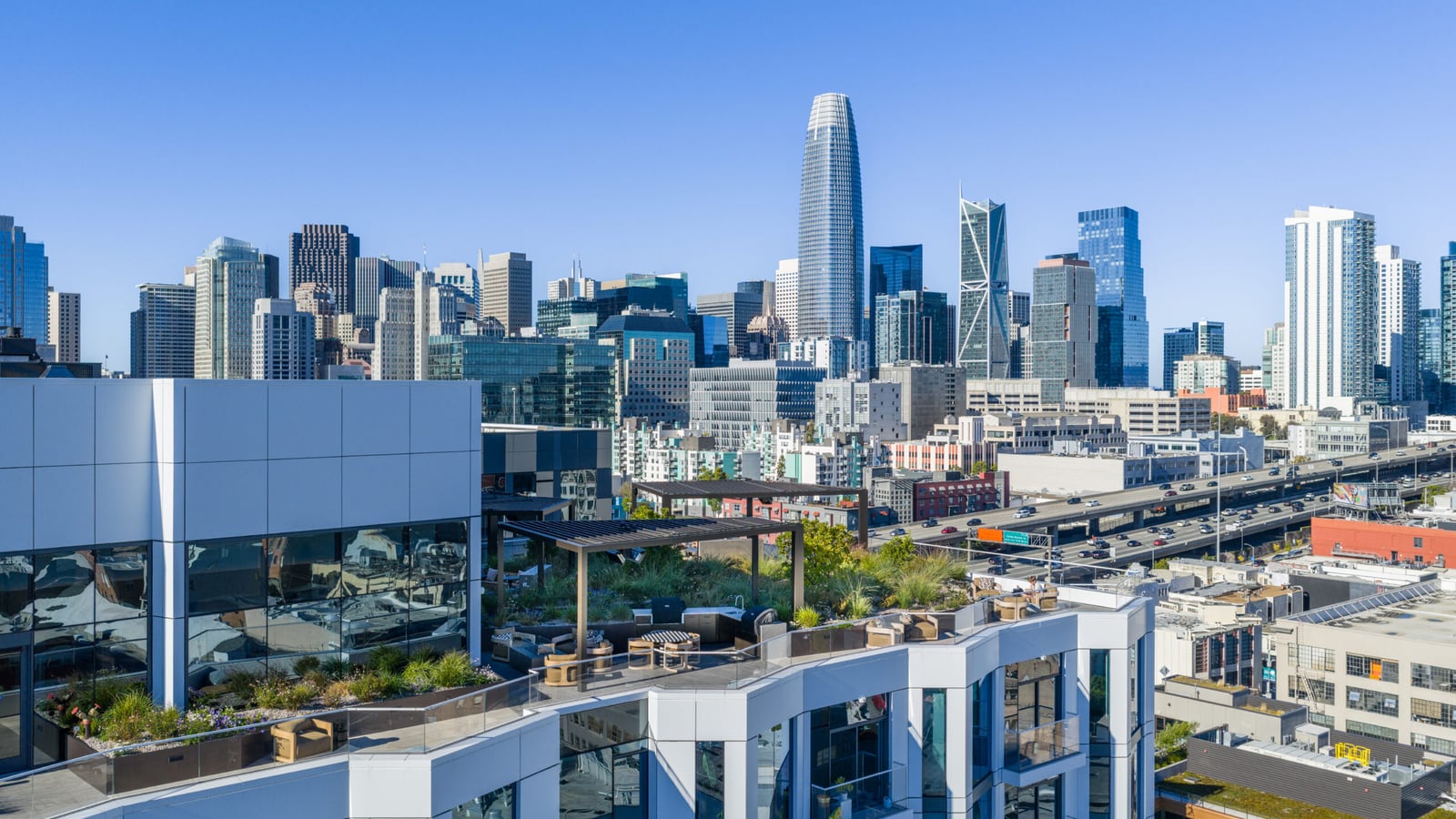 Photograph of a planted roof terrace with pergolas and seating on top of the building, with the downtown San Francisco skyline, including Salesforce Tower, behind it