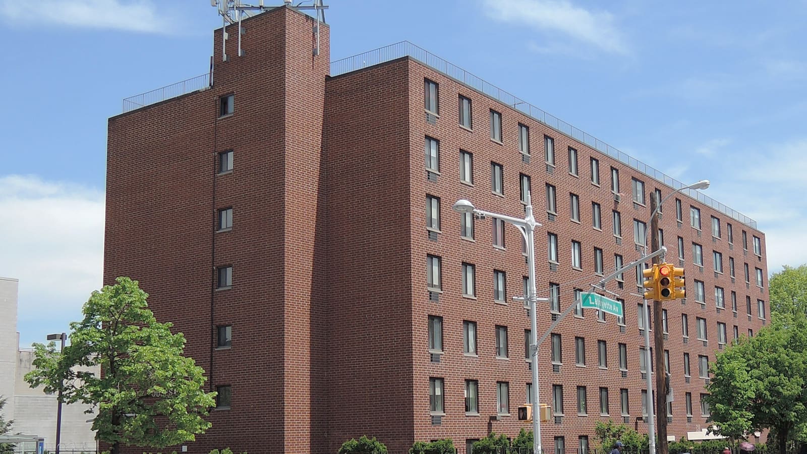 A seven-story red brick apartment house behind a traffic light and a Lafayette Av street sign, with trees at its base