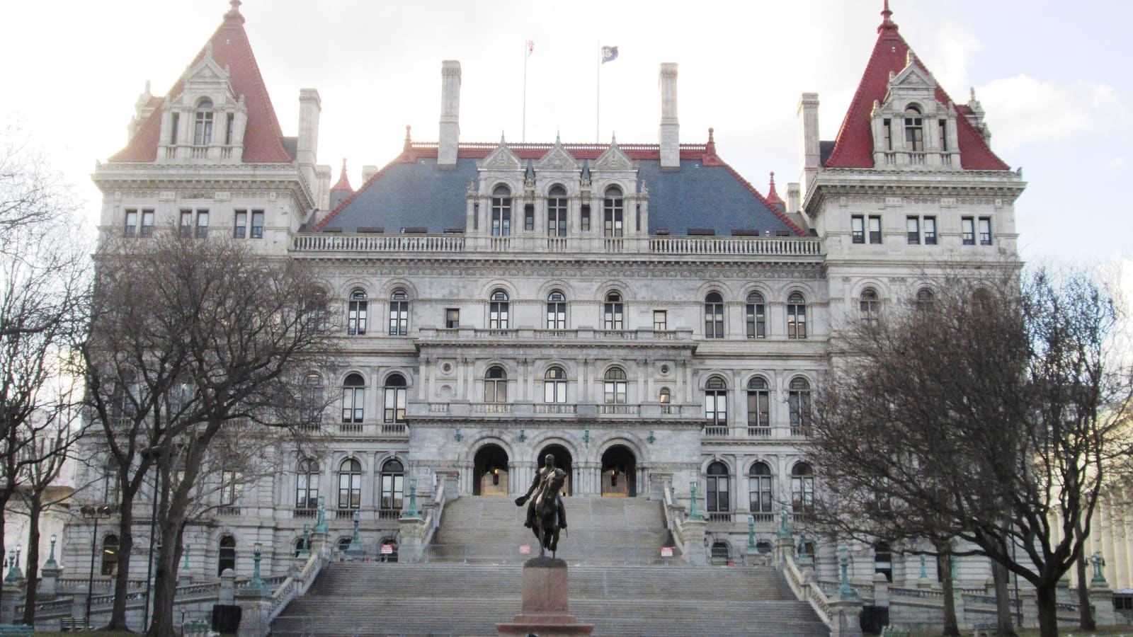 A large grey stone government building with red-roofed corner towers, a wide front staircase and bare trees on either side