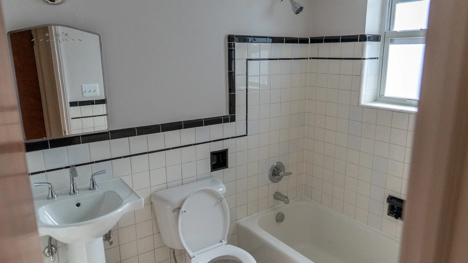 Photograph of a bathroom with white wall tile edged in black, a pedestal sink, a toilet, a tub and a window